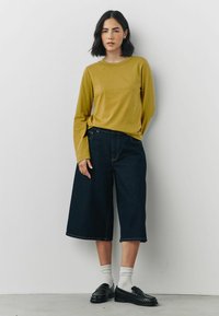 Langærmet sennepsgul bluse parret med brede, mørk denim culotter. Modellen bærer sorte loafers og hvide ribbede sokker, sat mod en ensfarvet baggrund.