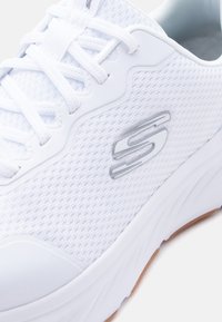 Λευκό αθλητικό παπούτσι με ύφασμα από δίχτυ, λευκά κορδόνια, γκρι εσωτερικό, χοντρή σόλα και μεταλλικό λογότυπο "S" στο πλάι.