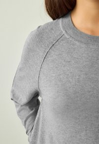 Grå sweater lavet af blødt materiale med ribbet halsudskæring, raglanærmer og glat tekstur, der viser afslappede designdetaljer.