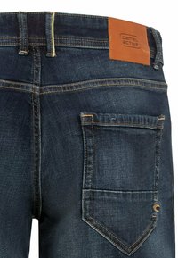 Mørkeblå denim jeans med en brun læderlap, der er mærket "camel active." De har synlige sømme og en baglomme med en spidseklap.