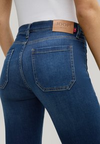 Jeans z denimu w ciemnoniebieskim kolorze, z wysokim stanem, delikatnym przetarciem i skórzaną naszywką z napisem "JOOP! JEANS" z tyłu.