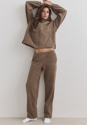 Mujer de pie con los brazos levantados, vistiendo chaqueta marrón de forro polar con cremallera y pantalones a juego, calcetines blancos, contra fondo de pared lisa y clara.