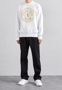 Vit sweatshirt med en guldfärgad cirkulär logotyp, svarta jeans och vita sneakers. Slät textur, rund halsringning samt ribbade mudd och fåll.