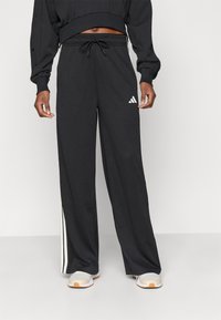 Čierne športové nohavice s textúrou z oka, elastickým pásom, šnúrkou a bielymi pruhmi po stranách. Na ľavom stehne sa nachádza logo Adidas.