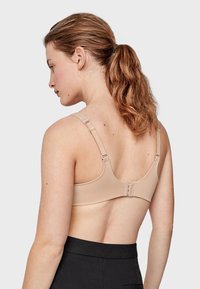 Soutien-gorge beige avec bretelles réglables et fermeture à crochets. Tissu lisse avec un design sans coutures. Modèle portant un pantalon noir, les cheveux en queue de cheval.