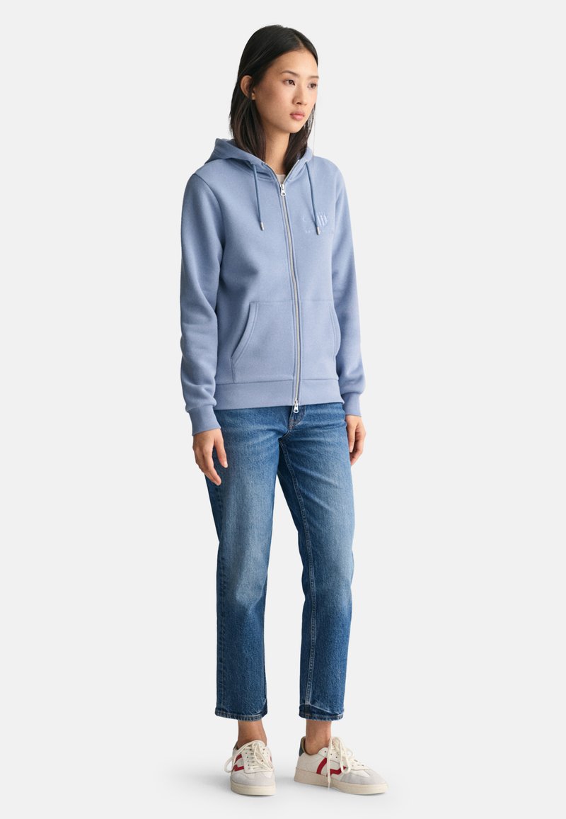 Lichtblauwe zip-up hoodie met een capuchon, vo zakken aan de voorkant en een trekkoord. Draag het met lichtblauwe jeans en witte sneakers. Glad textuur, casual ontwerp.