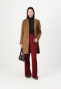 Brown suede coat with a tailored design, black turtleneck, pleated burgundy pants, and leopard-print shoes. Black handbag with straps.