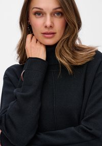 Kobieta z jasnobrązowymi włosami, nosząca czarny sweter z golfem, delikatnie opiera rękę na szyi, patrząc przed siebie na jednolitym tle.