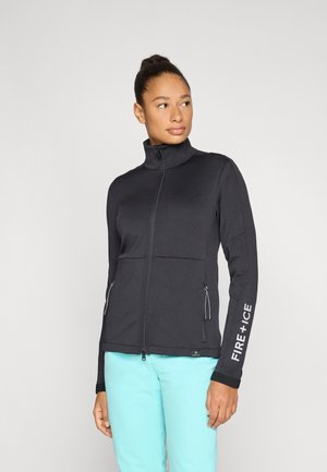 Woman with hair in a bun wearing a black zip-up jacket labeled "FIRE + ICE" on the sleeve and light blue pants, looking to the side.