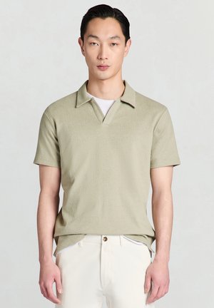 Mann trägt ein hellgrünes, strukturiertes kurzärmeliges Poloshirt über einem weißen Unterhemd und hellbeigen Hosen vor einfachem Hintergrund.