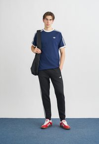 Tmavomodré športové tričko s bielymi pruhmi, čierne teplákové nohavice so stranovými pruhmi, červené tenisky a čierna taška na rameno.