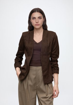 Jeune femme aux cheveux foncés portant un blazer marron, un haut marron foncé et un pantalon plissé beige taille haute, debout devant un fond blanc uni.
