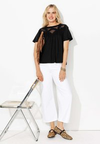 Femme blonde portant un top en dentelle noire, un pantalon large blanc, des ballerines imprimé léopard, tenant un sac en franges marron, se tenant à côté d'une chaise transparente.