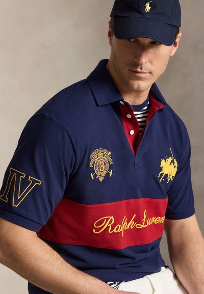 Άνδρας που φορά μπλε ναυτικό και κόκκινο πόλο Ralph Lauren με κεντημένα λογότυπα και μπλε ναυτικό καπέλο με κίτρινο λογότυπο, καθισμένος σε απλό φόντο.