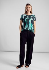 Top a maniche corte con un motivo astratto verde e nero, abbinato a pantaloni neri larghi e sandali neri piatti. Design minimalista.