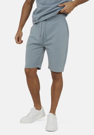 Mężczyzna regulujący niebieskie, teksturowane spodenki ze ściągaczem, ubrany w białe trampki, stojący na tle jednolitej powierzchni.