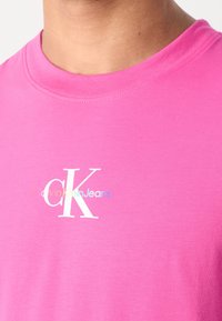 Růžové bavlněné tričko s kulat�ým výstřihem. Zdobeno bílým logem s nápisem "CalvinKleinJeans" v jemných barvách pod emblémem "CK".