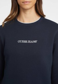 Felpa navy con scollo girocollo a costine, caratterizzata dal logo ricamato "GUESS JEANS" bianco sul petto. Tessuto liscio, vestibilità casual.