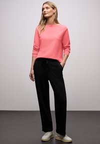 Kobieta stojąca z rękami w kieszeniach czarnych spodni, ubrana w różową bluzkę z długim rękawem i białe trampki na szarym tle.