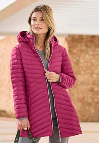 Frau trägt eine lange, rosafarbene, gesteppte Jacke mit Kapuze, die teilweise zugezippt ist, hält einen dunkelgrünen Taschenriemen und steht im Freien.