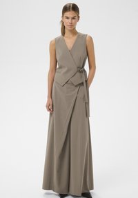 Kvinde stående iført taupe ærmeløs slå-om vest og matchende vidde bukser med bælte, foran en ensartet lys baggrund.