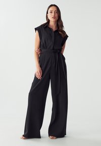 Zwarte jumpsuit met een kraag, korte mouwen en een gestrikt riem; gemaakt van een gladde stof; wijde pijpen en minimalistische stijl.