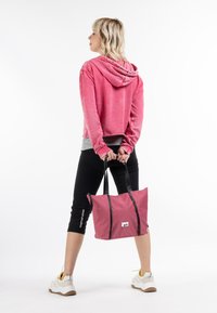 Borsa a tracolla rosa con manici neri, realizzata in tessuto resistente. Presenta una chiusura con zip e una patch con logo. Modello che indossa un felpa rosa e leggings neri.