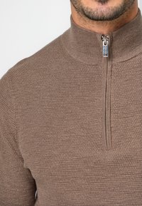 Brauner Strickpullover mit hohem Kragen und halbem Reißverschluss. Strukturierter Stoff mit einem dezenten Muster, entworfen für Wärme und Komfort.