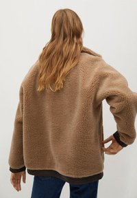 Brązowa kurtka z futra sztucznego o luźnym kroju, w oversize'owym kształcie i fakturowanej powierzchni. Posiada kołnierz szalowy oraz kontrastowe ciemniejsze mankiety.