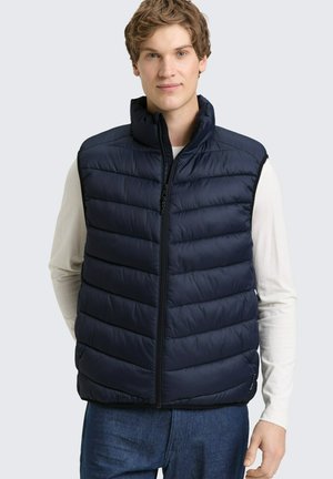 Junger Mann trägt eine marineblaue gesteppte ärmellose Weste über einem weißen Langarmshirt und blauen Jeans, steht vor einem einfarbigen Hintergrund.