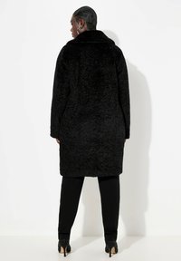Abrigo de piel sintética negra con una textura suave, con un cuello oversize y mangas largas. Se lleva con estilo sobre pantalones negros, llegando hasta la mitad del muslo.