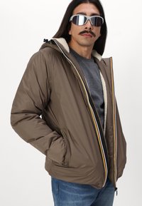 Giacca isolante marrone con chiusura a zip, caratterizzata da una fodera interna a contrasto, tasche laterali e accenti a righe multicolori sulla zip.