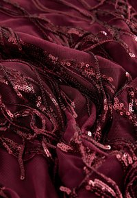 Burgundy mesh fabric with flowing strands of sequins, featuring a textured surface and a cascading design, reflecting light with shimmering accents.