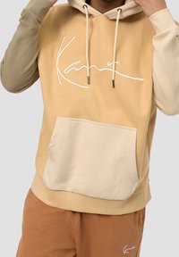 Huvtröja med beige och ljusbruna färgblock, framficka och broderad signatur. Blandad bomullstyg och justerbara snörningar.