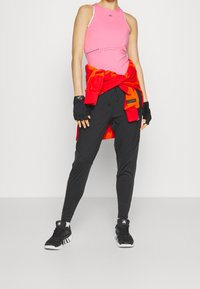 Różowy sportowy top na ramiączkach z okrągłym dekoltem, czarne spodnie dresowe, czerwona bluza zawiązana w talii oraz czarne sportowe buty. Teksturowane rękawiczki.