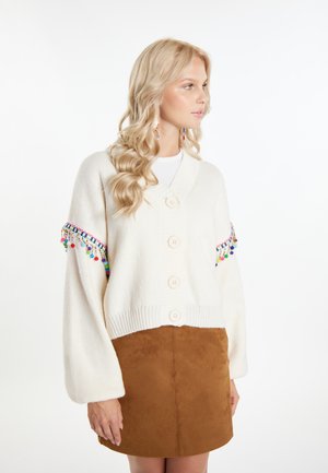 Witte gebreide cardigan met grote knopen, brede mouwen en kleurrijke pompons langs de manchetten. Gecombineerd met een bruine suède rok.