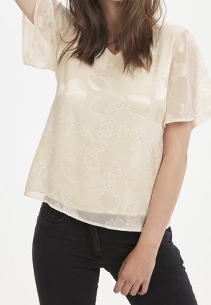 Frau trägt eine durchsichtige cremefarbene Bluse mit floralem und Muschelmuster, kombiniert mit schwarzen Hosen, posiert mit einem erhobenen Arm.