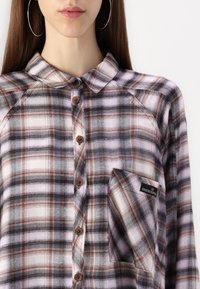 Ruutukuvioinen flanellipaita vaaleanpunaisen, ruskean ja harmaan sävyissä, jossa on nappikaulus, rintatasku ja puunapit.