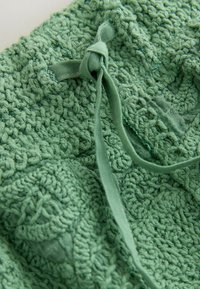 Tissu tricoté vert avec motif texturé et détail de noeud tissé, noué lâchement sur un fond clair.