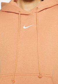 Persikofärgad hoodie med framfickan av typ kanguro, vit Nike-logotyp, snoddar med metalldelar och en strukturerad tygfinish.