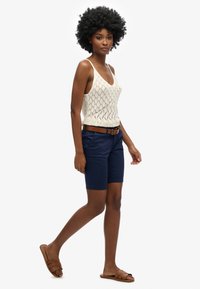 Modella con un top in cotone all'uncinetto color crema, shorts blu scuro e sandali scivolosi marroni. L'outfit presenta un design aderente con una trama tessuta e una cintura.