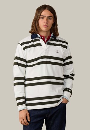 Joven con cabello largo y castaño que lleva una camiseta de rugby blanca con rayas horizontales oscuras y vaqueros oscuros, de pie con una mano en el bolsillo.