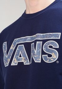 Sweat-shirt bleu marine arborant un grand logo "VANS" avec un design de paysage bleu, incluant des palmiers et des accents de soleil, imprimé sur le devant.