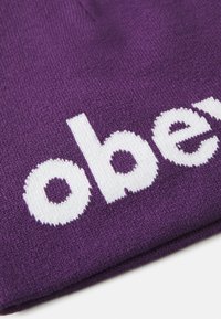 Violeta adīta cepure ar baltu izšūtu tekstu. Maiga tekstūra ar tuvplānu, kas izceļ auduma aušanas rakstu un burtiem.