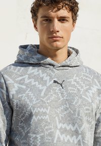 Noor mees lokkis juustega, kandes helehallit mustriga kapuutsi, millel on must Puma logo, ühetaolise heleda tausta ees.