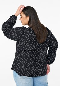 Blusa nera con un motivo geometrico bianco, maniche lunghe con polsini elasticizzati e vestibilità comoda, abbinata a jeans blu chiaro.