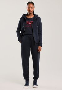 Moteris stovinti, vilkinti tamsiai mėlyną užtraukiamą džemperį su gobtuvu, derančius sportinius kelnes, raudoną marškinėlį su grafiniu atspaudu ir tamsiai mėlynas sportines basutes, stovinti priešais paprastą foną.