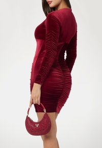 Vestido de terciopelo rojo con mangas largas, textura recogida y diseño ajustado. Combinado con un bolso rojo brillante de forma curvada.