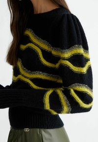 Femme portant un pull noir en maille avec des rayures ondulées jaunes et des accents métalliques, associée à un pantalon vert et de longs cheveux bruns.