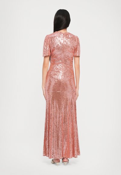 Vrouw met lang, donker haar die een floor-length, korte mouwen rosegouden pailletten jurk en beige hakken draagt, staand met haar rug naar de camera op een witte achtergrond.